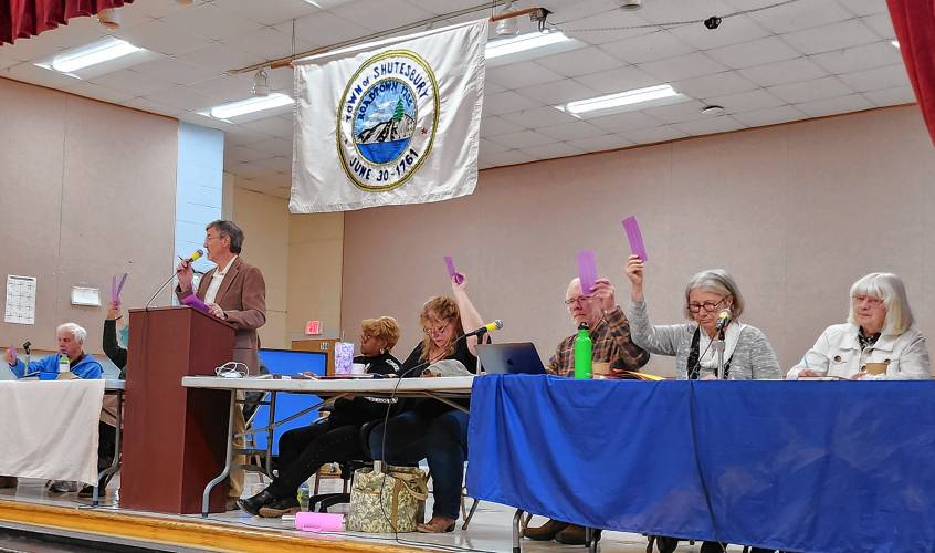 Members of Shutesbury’s Finance Committee and Selectboard are seated on the stage during Saturday’s Annual Town Meeting at Shutesbury Elementary School. Moderator Paul Lyons is at the podium.