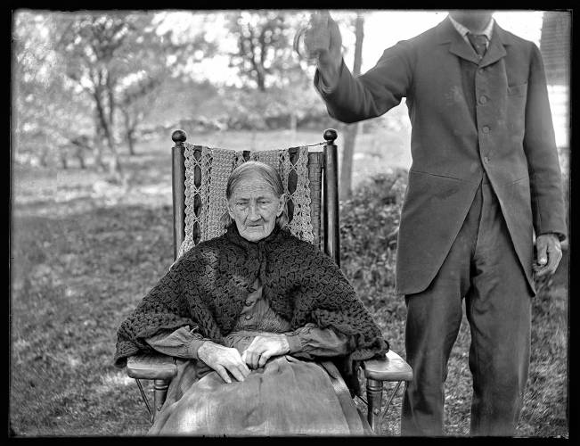In an image from the 1890s, an elderly woman sits for a portrait while a man holds something over her. 