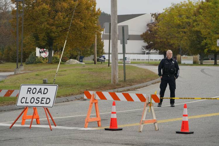 A police officer stands at a road closure near a bowling alley, seen in background, Thursday, Oct. 26, 2023, in Lewiston, Maine. The site is one of Wednesday's two mass shootings in the city. Residents have been ordered to shelter in place as police continue to search for the suspect. (AP Photo/Robert F. Bukaty)